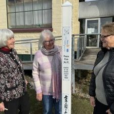 Three women looking at a white pole featuring a dove bearing an olive branch and the insignia of the Rotary Club of Tasmania and the words "A Century of Service Since 1924". 100 years. Underneath are the words Peace Pole Number 51. Donated by the Rotary Club of West Tamar.